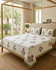 A bed dressed in a white cotton sheet and matching pillowcases with a green and brown botanical print, styled in a modern bedroom with a wooden canopy bed, large windows, and a view of palm trees outside.