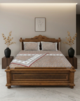 A fully made bed with a patterned beige and orange bedding set, set against a dark carved wooden bed frame. The bed is flanked by two black side tables with dried plants in black vases, set against a warm beige wall with a minimalist art print.