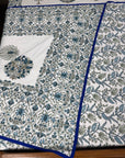 A folded white fabric textile with an intricate blue and green floral block print pattern and a solid blue border, displayed on a wooden surface.