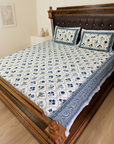 A bed dressed in a white quilted bedspread and matching pillowcases with a blue floral and paisley pattern, set in a bedroom with a dark brown tufted wooden headboard and light wood flooring.