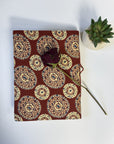 A fabric-covered notebook or journal with a repeating circular medallion pattern in rust red, cream, and blue, styled on a white surface with a dried rose resting on top and a small potted succulent nearby.