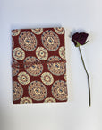 A small fabric-covered notebook or journal with a red-burgundy cover and cream, beige, and blue circular medallion pattern, shown on a white background next to a single dark purple dried rose stem.