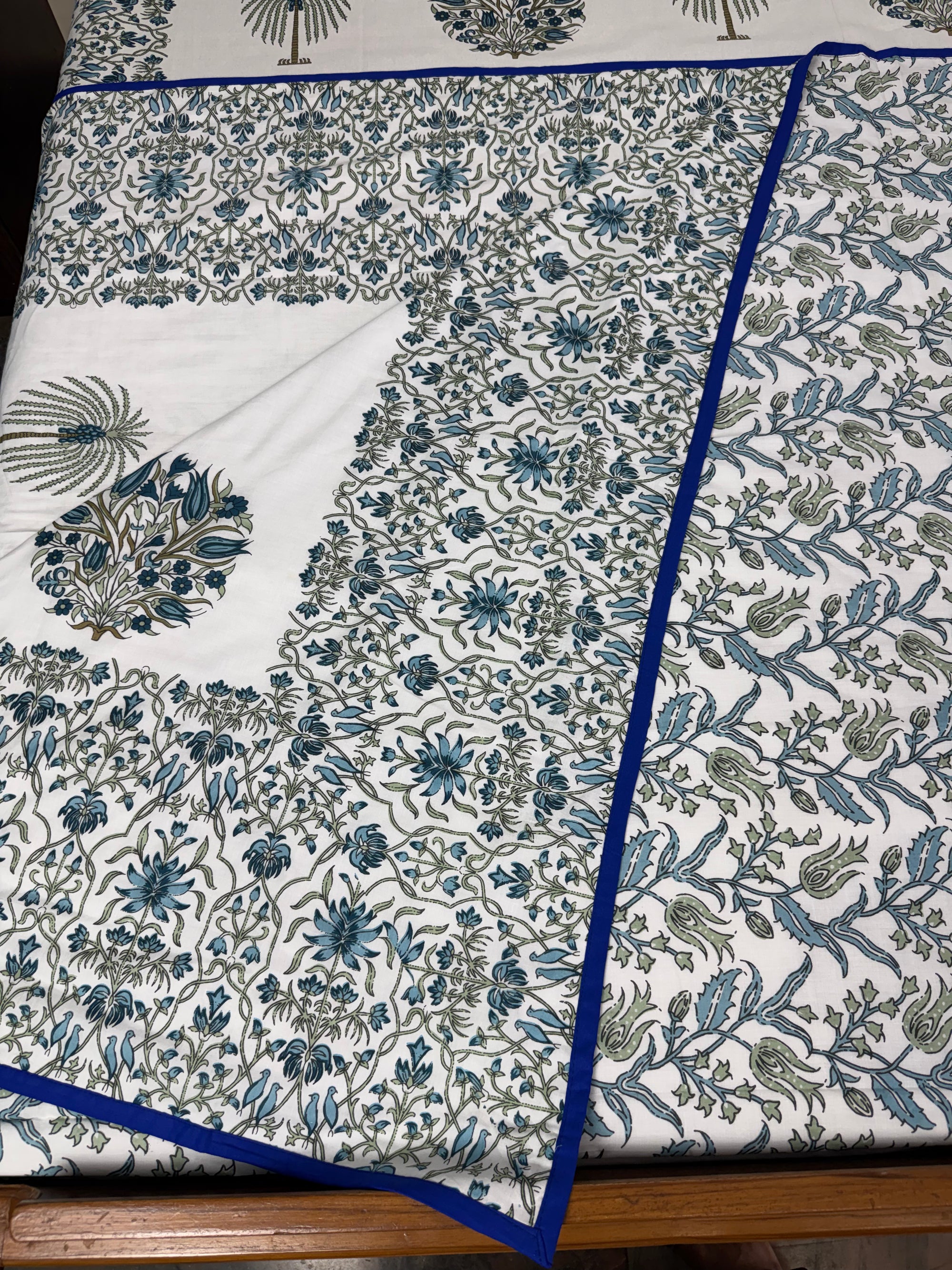 A folded white fabric textile with an intricate blue and green floral block print pattern and a solid blue border, displayed on a wooden surface.