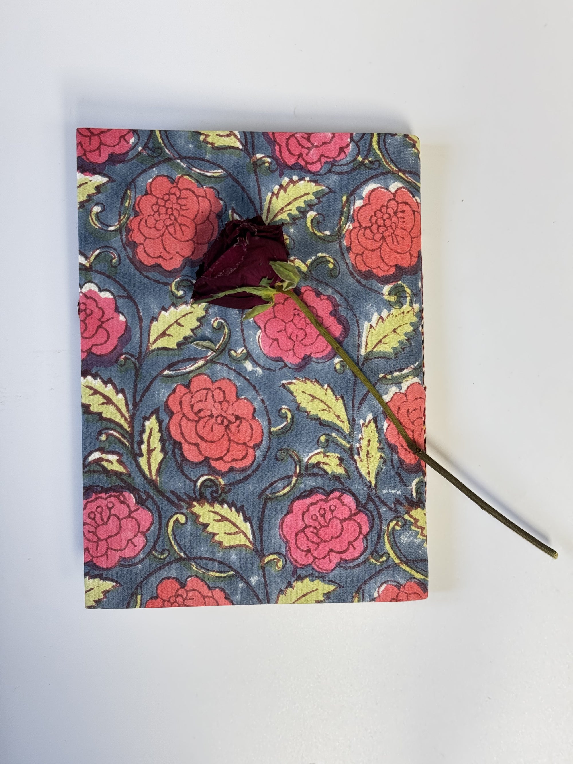 A square notebook or journal with a vintage floral pattern in pink, red, and yellow on a dark blue-gray background, decorated with a single dark red rose pressed on top, set against a plain white background.