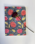 A square notebook or journal with a vintage floral pattern in pink, red, and yellow on a dark blue-gray background, decorated with a single dark red rose pressed on top, set against a plain white background.