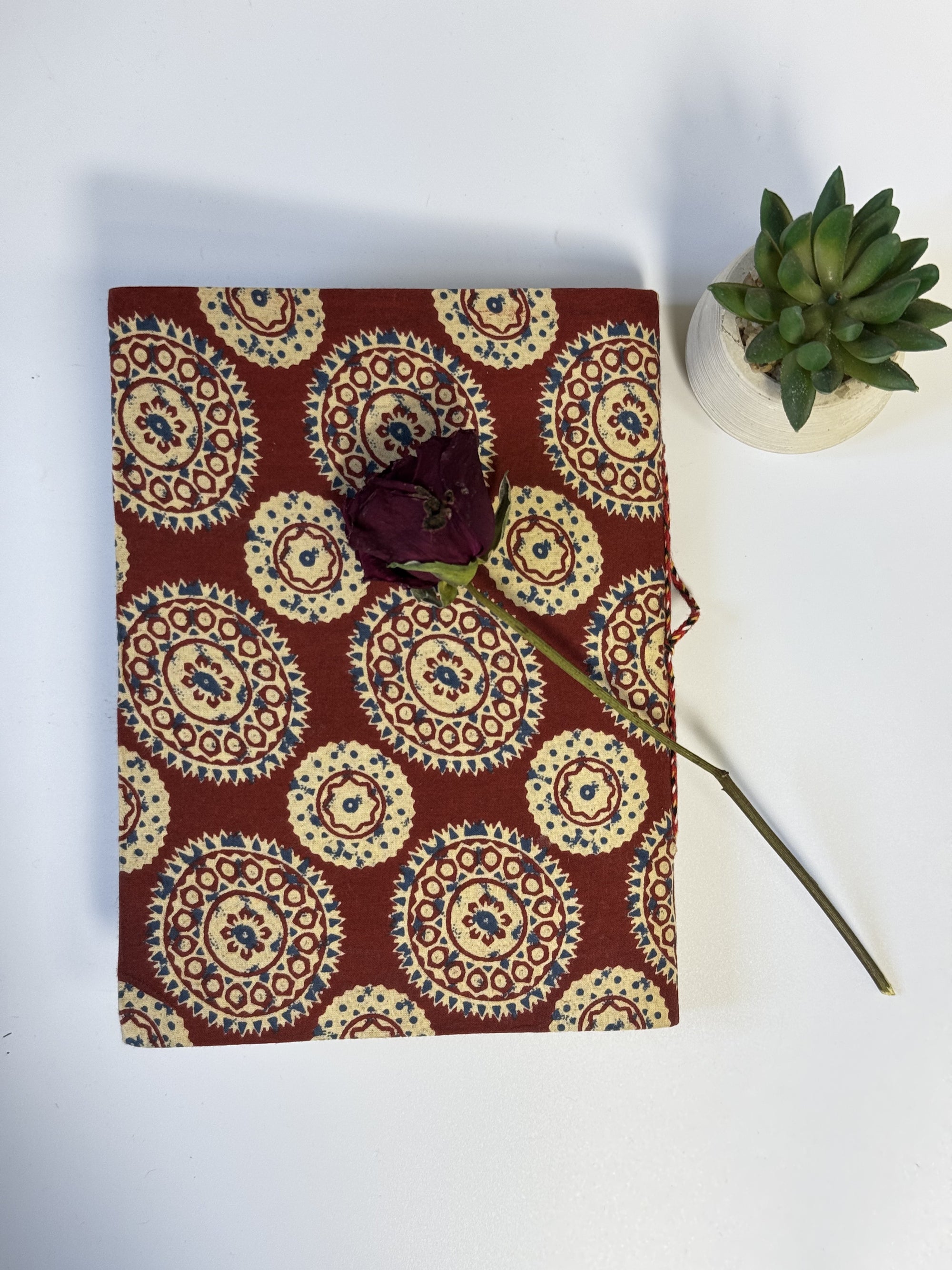 A fabric-covered notebook or journal with a repeating circular medallion pattern in rust red, cream, and blue, styled on a white surface with a dried rose resting on top and a small potted succulent nearby.