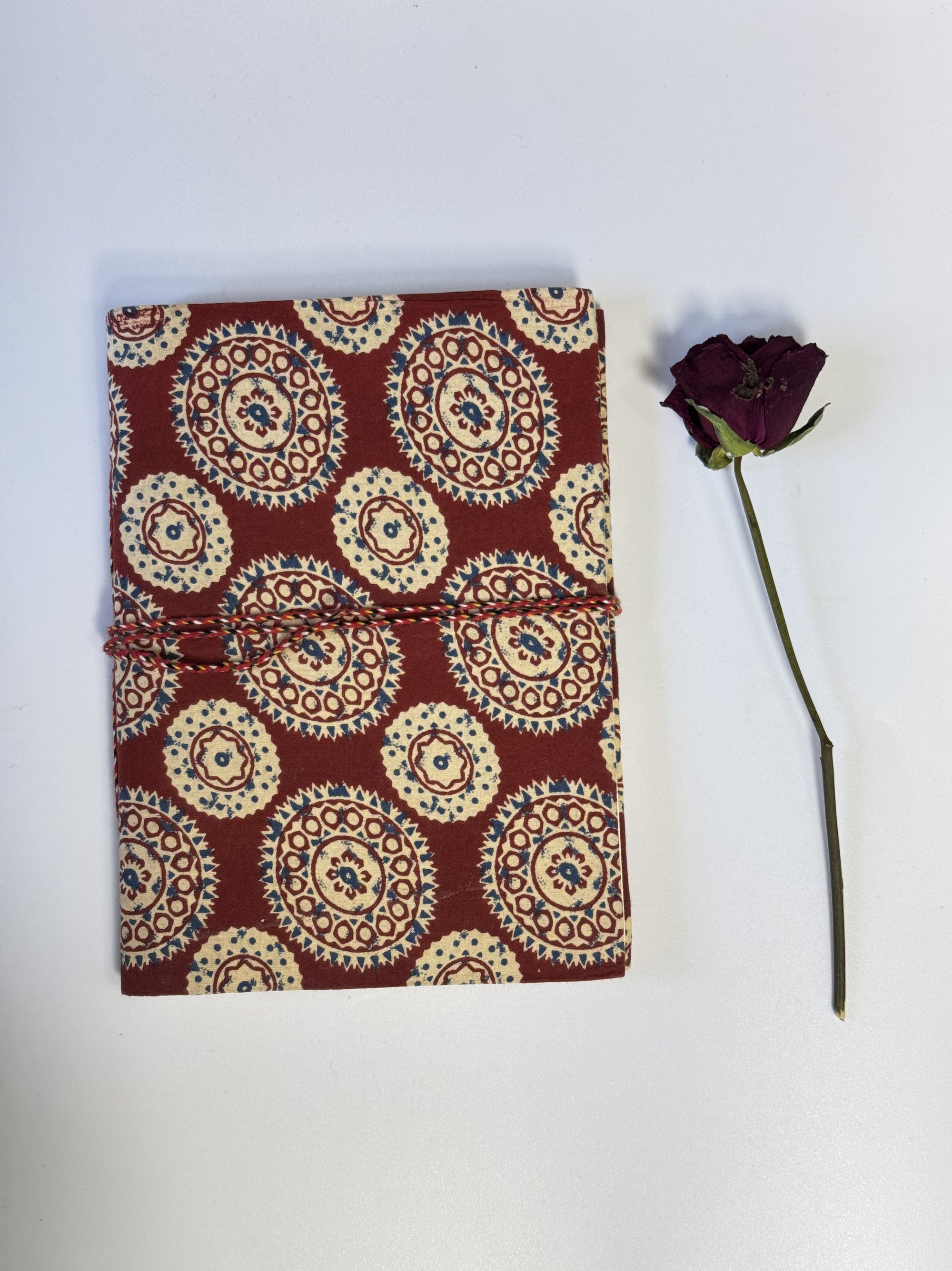 A small fabric-covered notebook or journal with a red-burgundy cover and cream, beige, and blue circular medallion pattern, shown on a white background next to a single dark purple dried rose stem.