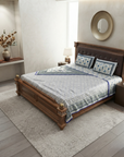 A styled bedroom scene showing a wooden bed dressed with a blue and white patterned quilt and matching pillows, placed in a real interior with a rug, wall decor, and bedside lamp.