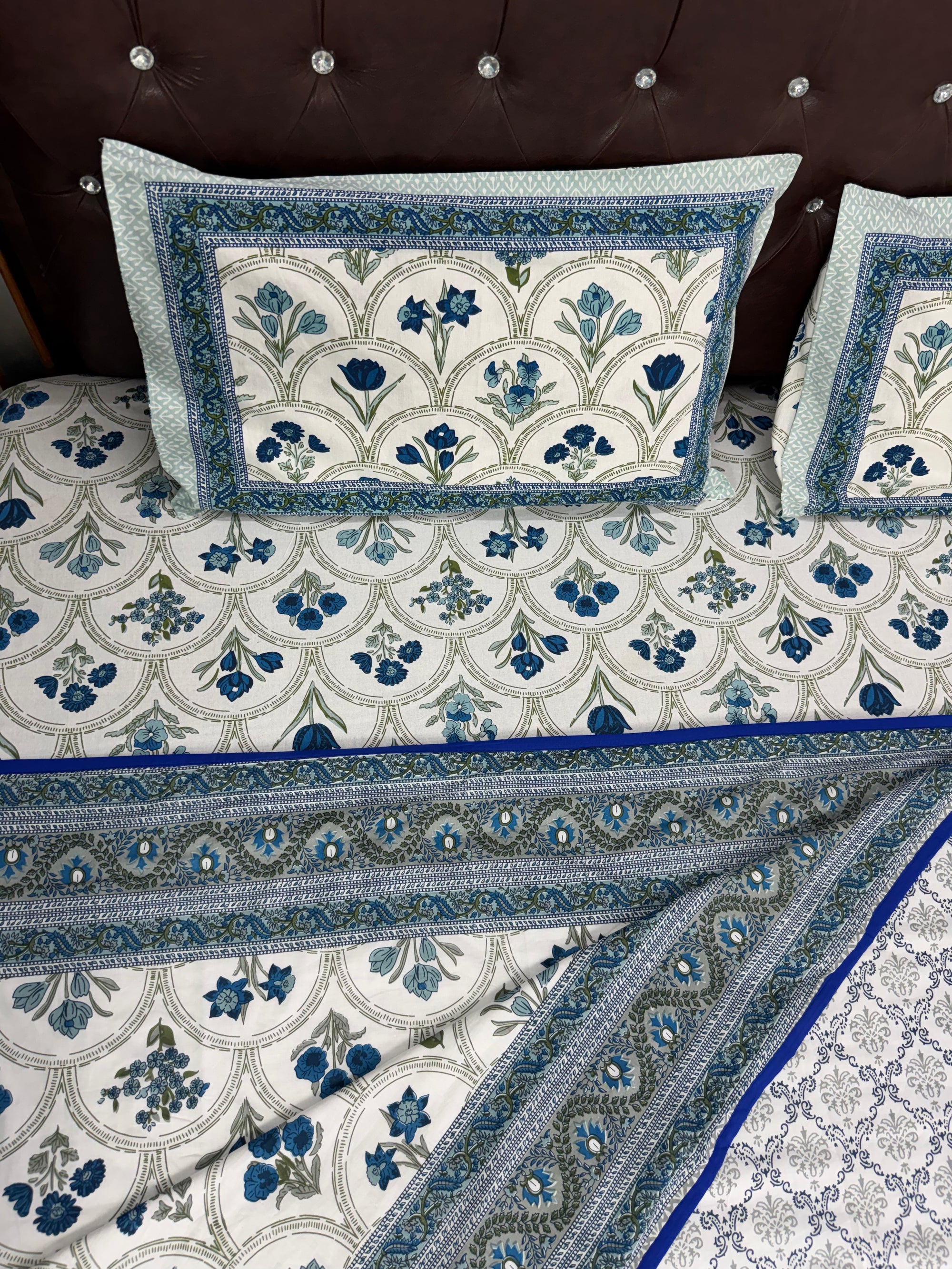 A blue and white patterned cotton bedspread set with matching pillowcases, styled on a bed with a dark brown tufted headboard.