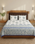 A neatly made bed in a styled bedroom setting, featuring a white and blue patterned cotton sheet set with matching pillowcases and a decorative blue border. The bed has a brown leather and wood headboard, flanked by wooden bedside tables with decorative items.