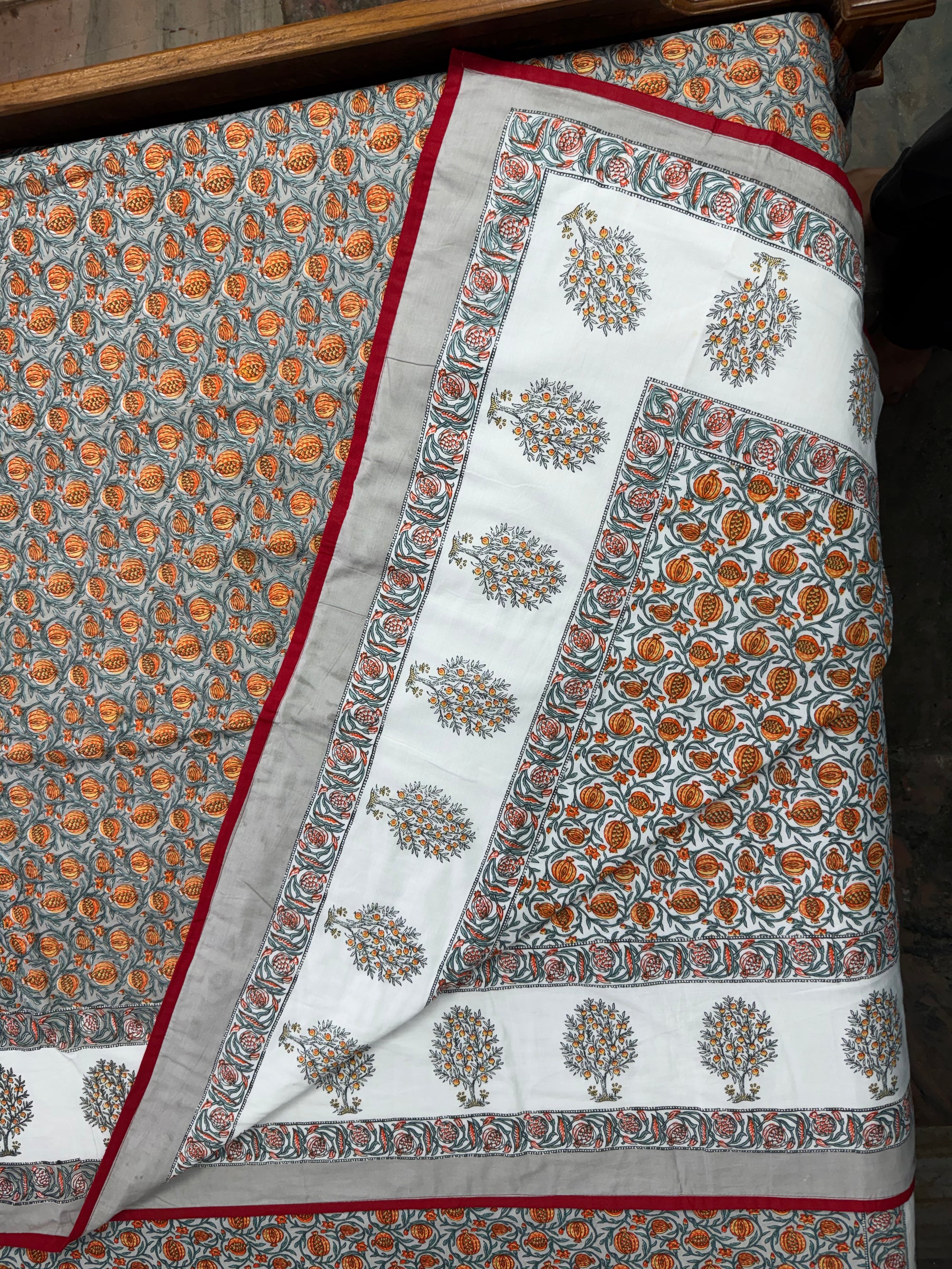 A folded and draped block-printed cotton textile, likely a quilt or comforter, featuring a white central panel with orange and gray floral motifs, a gray border with red piping, and a matching patterned section.