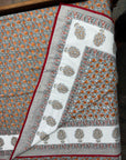 A folded and draped block-printed cotton textile, likely a quilt or comforter, featuring a white central panel with orange and gray floral motifs, a gray border with red piping, and a matching patterned section.