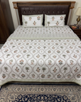A bed dressed in a white patterned quilted sheet set with matching pillowcases, featuring a repeating geometric and palm tree motif. The bed has a dark brown tufted wooden headboard, a bedside lamp, and a patterned rug on the floor.