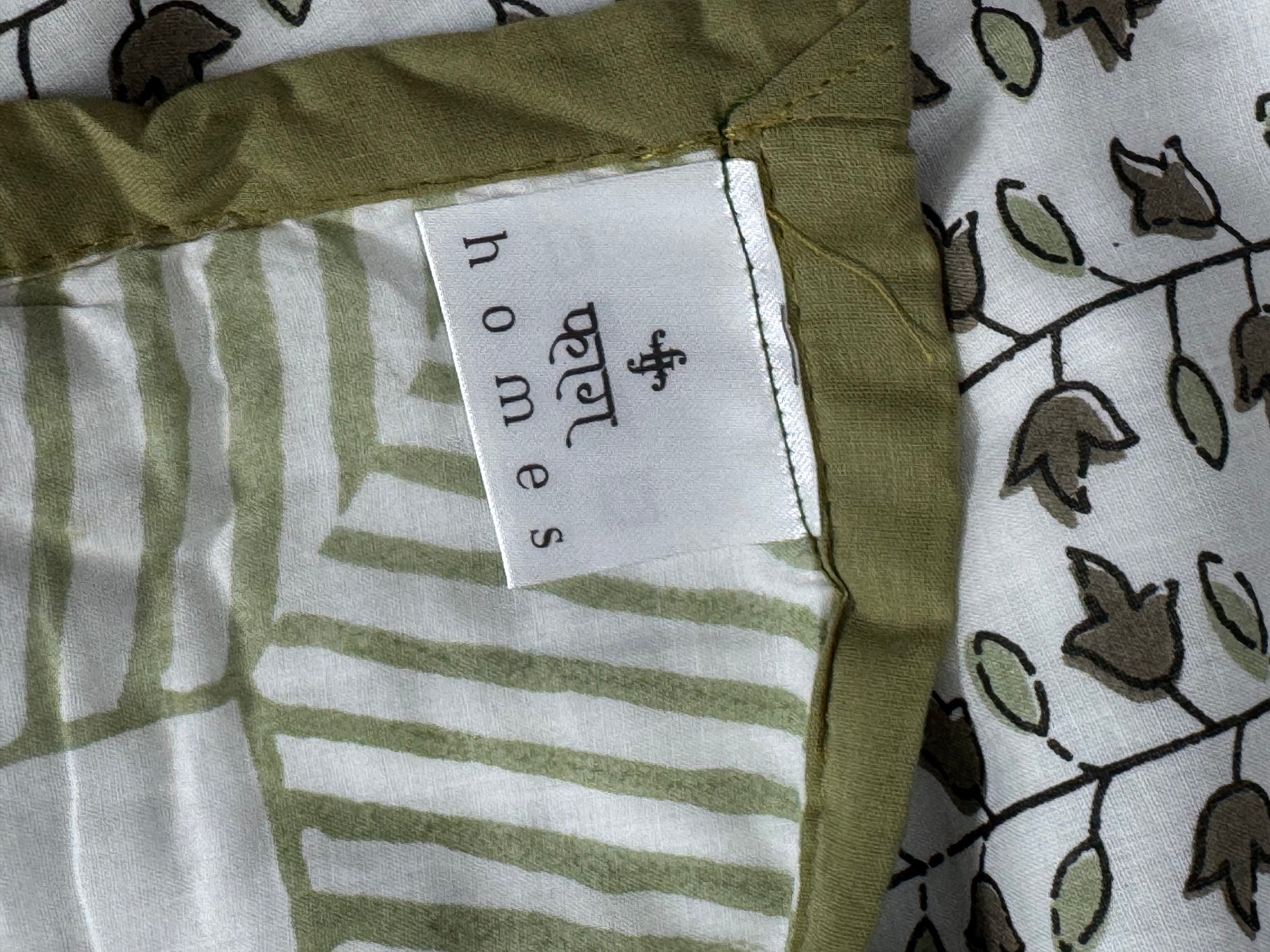 Close-up of a fabric product showing a white sewn-in label that reads 'homes' with a small logo, alongside an olive green border and a white patterned textile with green and brown leaf motifs.