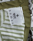 Close-up of a fabric product showing a white sewn-in label that reads 'homes' with a small logo, alongside an olive green border and a white patterned textile with green and brown leaf motifs.