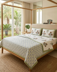 A neatly made bed in a bright bedroom featuring a white quilted cover with a blue geometric pattern and green border, matching pillow shams with a tree motif, and a wooden canopy bed frame. Large windows and curtains reveal a tropical garden outside.