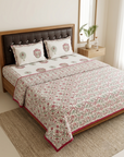 A neatly made bed in a bedroom setting featuring a cream-colored cotton quilt set with a pink and green floral pattern, matching pillowcases, and a solid pink border. The bed is styled with a dark brown tufted headboard, a wooden nightstand, and a woven rug.