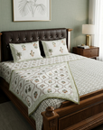 A neatly made bed in a styled bedroom, featuring a cream-colored quilted blanket and matching pillow shams with green and brown botanical palm tree patterns, set against a dark brown tufted headboard and a wooden nightstand.